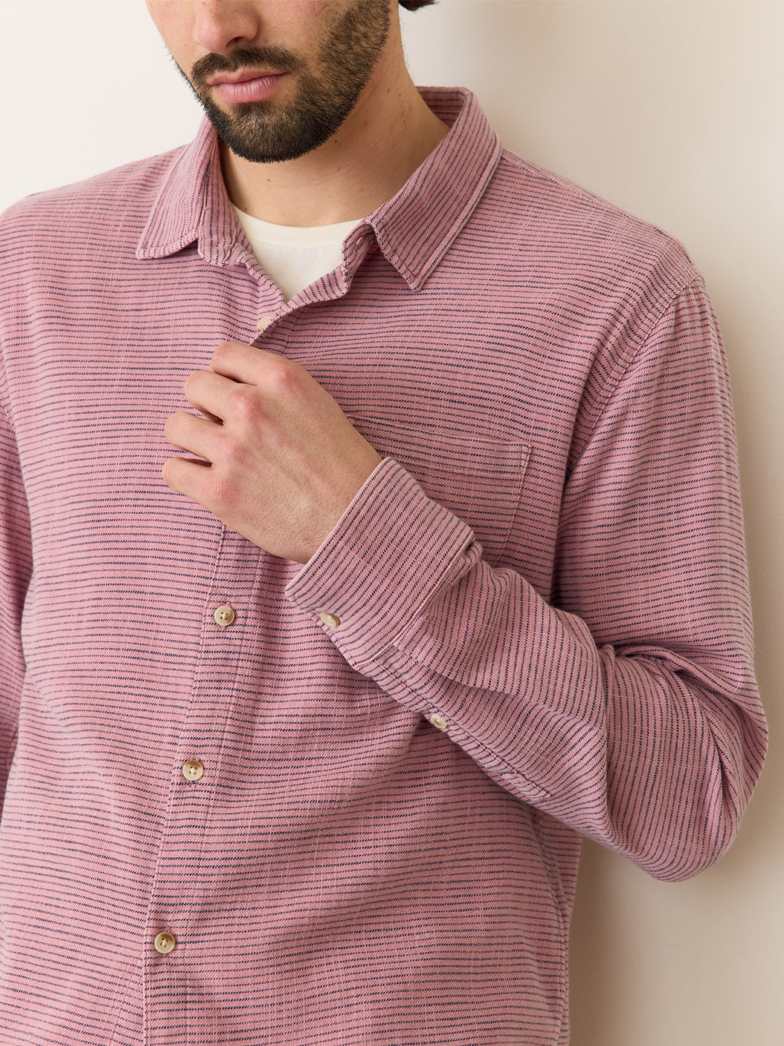 A person in a Marine Layer Stretch Selvage Long Sleeve Shirt with red and white stripes adjusts the top button with their right hand, highlighting its collar and buttons. The background is plain and neutral.