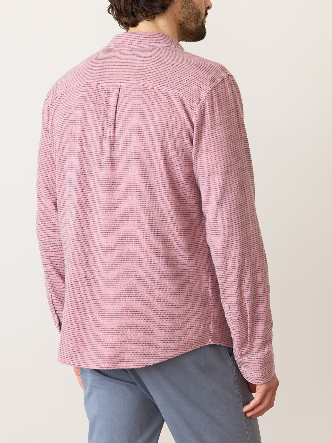 A person wearing the Marine Layer Stretch Selvage Long Sleeve Shirt in pink and white pinstripes, paired with gray pants, stands with their back to the camera. The shirt complements their short hair against a plain light background.