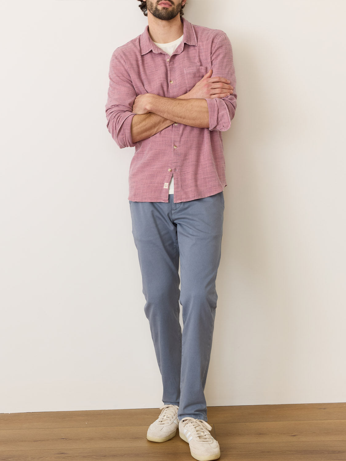 Standing against a beige wall, someone wears a pink Marine Layer Stretch Selvage Long Sleeve Shirt and blue pants. Paired with white sneakers and one leg bent, they give off an easygoing vibe while the wooden floor enhances their casual attire with its natural charm.