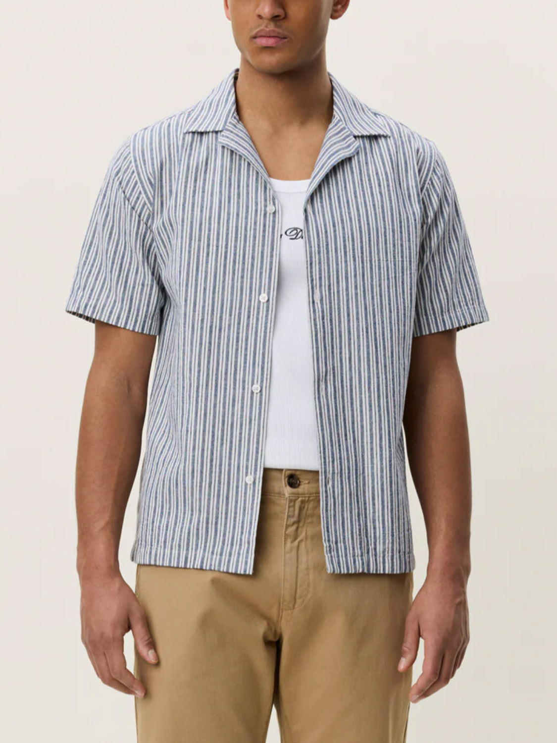 A person wears the Les Deux Stripe Cotton - Linen SS Shirt over a white ribbed tank with tan pants, standing against a plain light background.