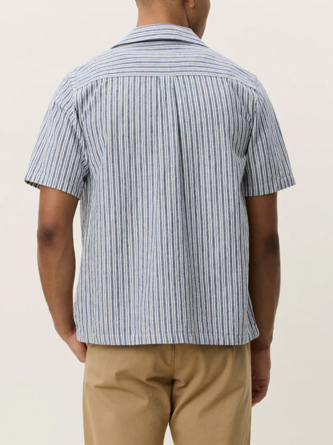A person stands facing away, wearing the Les Deux Stripe Cotton-Linen SS Shirt (short-sleeve, blue-and-white stripes) with light brown pants against a plain light background.