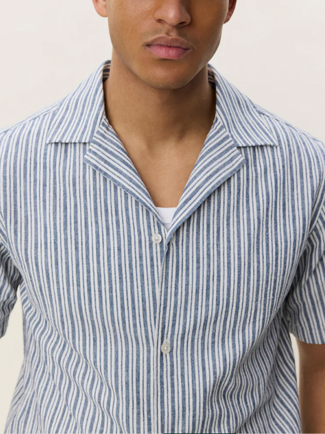 A person models the Les Deux Stripe Cotton - Linen SS Shirt, featuring blue and white vertical stripes and short sleeves, layered over a white undershirt against a plain light background, shown from shoulders to mid-torso.