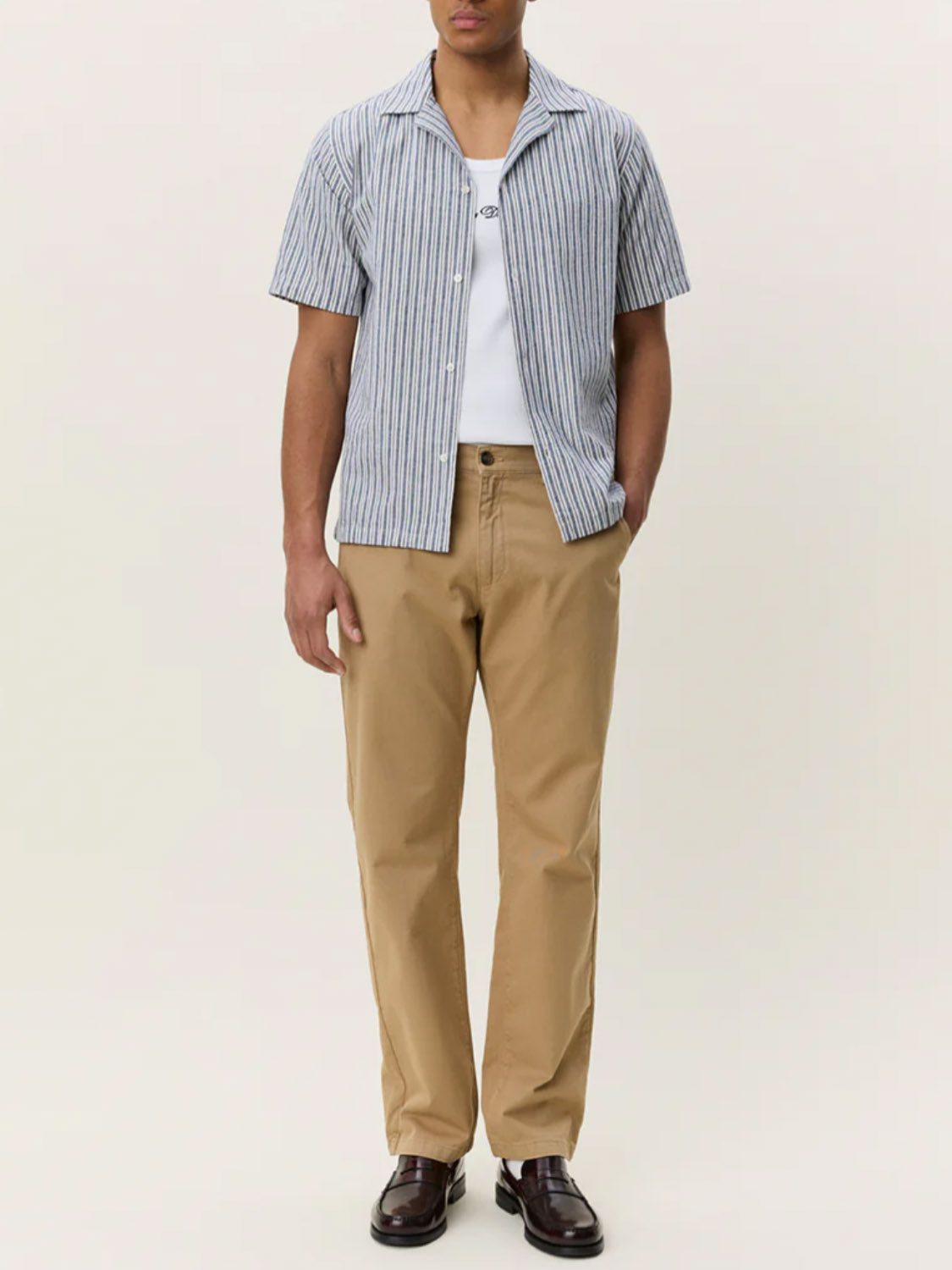 A man, hands in pockets and face partly hidden, models the Les Deux Stripe Cotton-Linen SS Shirt over a white tee with khaki pants and brown loafers against a plain background.