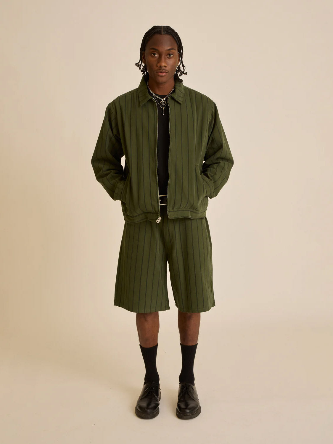 A person stands against a plain background wearing an Obey Stripe Denim Short set, featuring a matching green pinstripe jacket and baggy fit shorts with a black shirt, socks, and shoes. With hands casually in the jackets coin pockets, they gaze confidently at the camera.