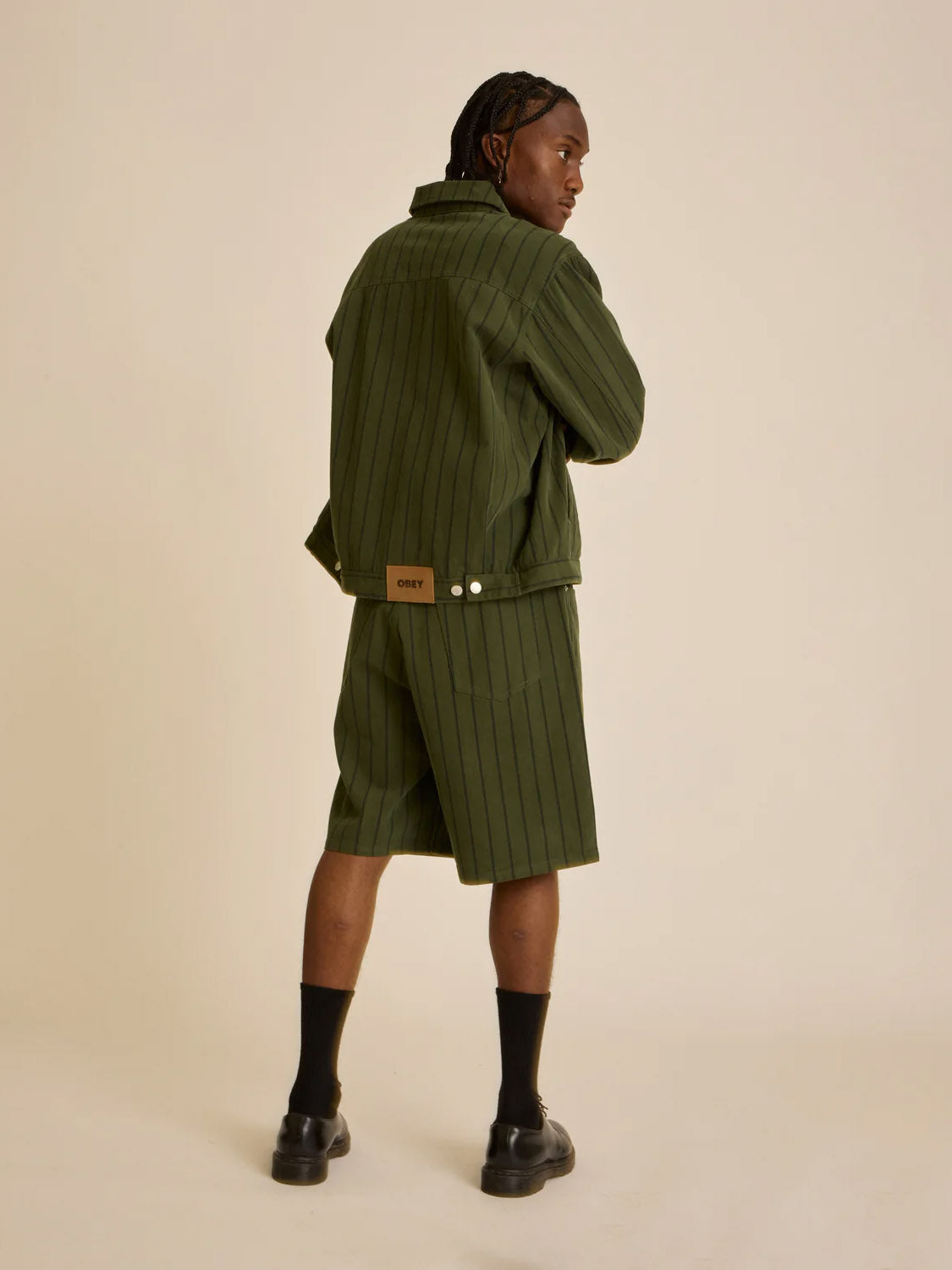 An individual in an Obey Stripe Denim Short and matching green pinstriped jacket, paired with black socks and shoes, poses with their back turned and head slightly to the side against a plain background.