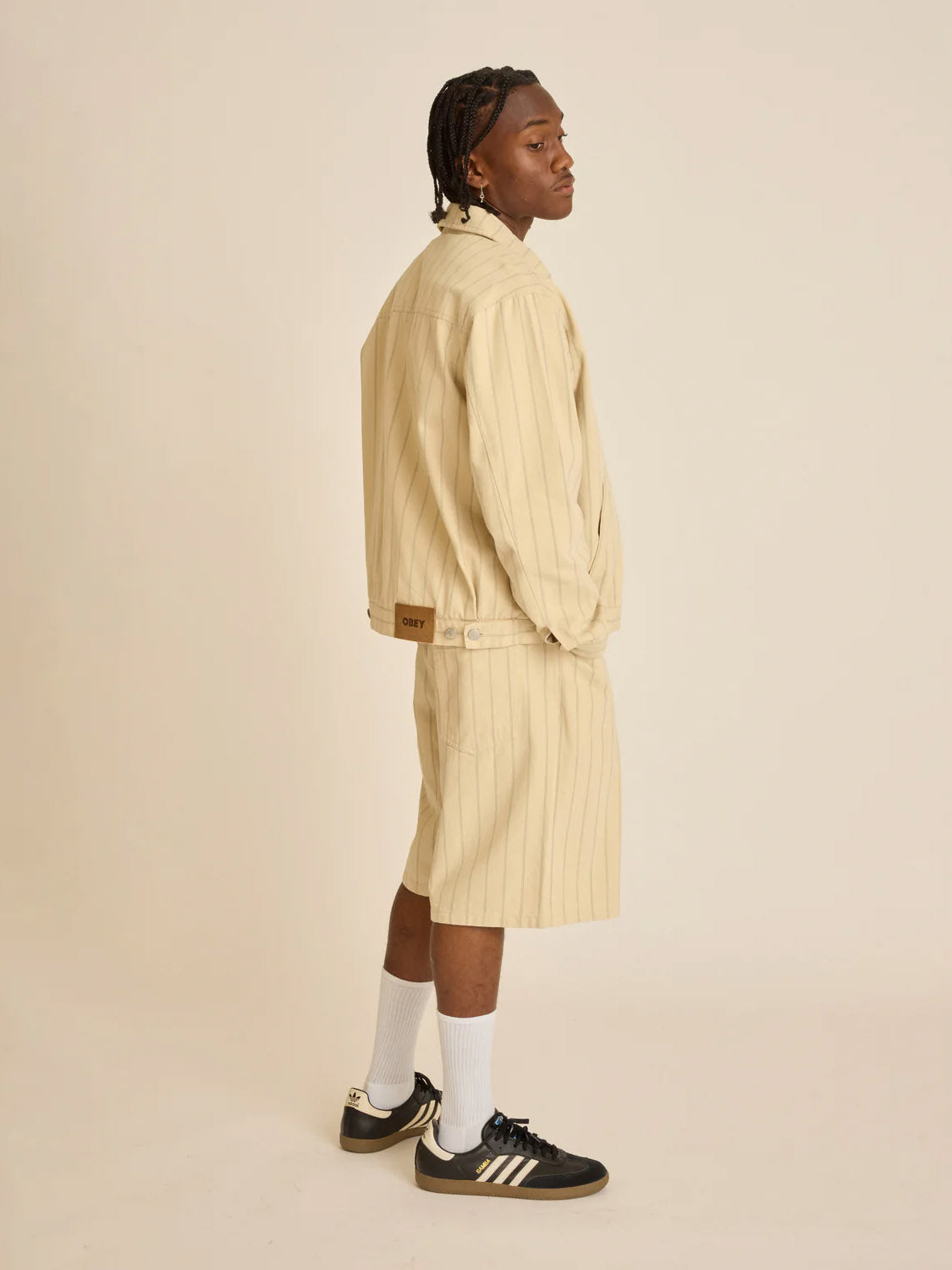 Wearing the Obey Stripe Denim Short and a matching light beige pinstripe jacket, a person poses sideways with braided hair. The outfit features a practical coin pocket, paired with white socks and black sneakers with white stripes on an off-white background.