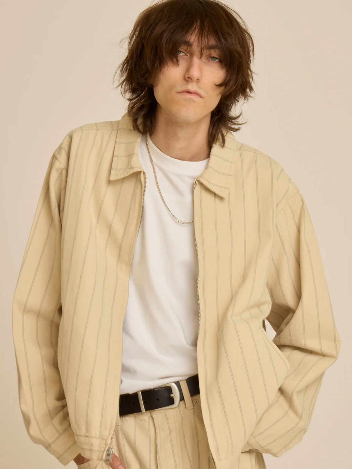 A person with shaggy hair models the Obey Stripe Zip Jacket, light beige and relaxed-fit, over a white t-shirt. They wear a necklace and gaze neutrally at the camera against a plain backdrop.