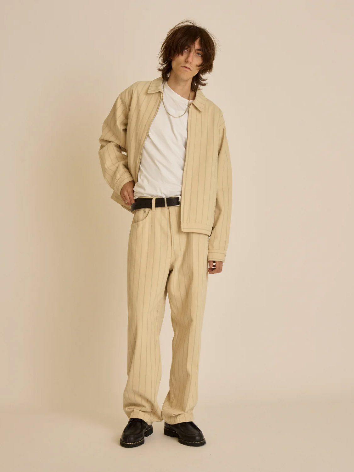 Someone is sporting a relaxed fit with an oversized light beige pinstripe Obey Stripe Zip Jacket and matching pants, complemented by a white T-shirt and black belt. Their medium-length hair adds to the casual vibe, while black shoes complete the look against a plain, light background.