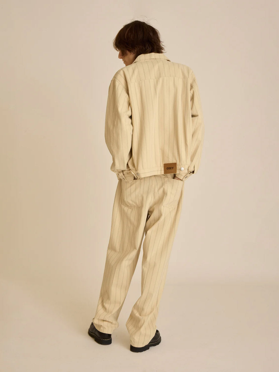 A person faces away from the camera, highlighting the Obey Stripe Zip Jacket—a beige, pinstriped pantsuit in a relaxed fit by Obey. The ensemble includes wide-legged pants and is paired with black shoes on a neutral background.