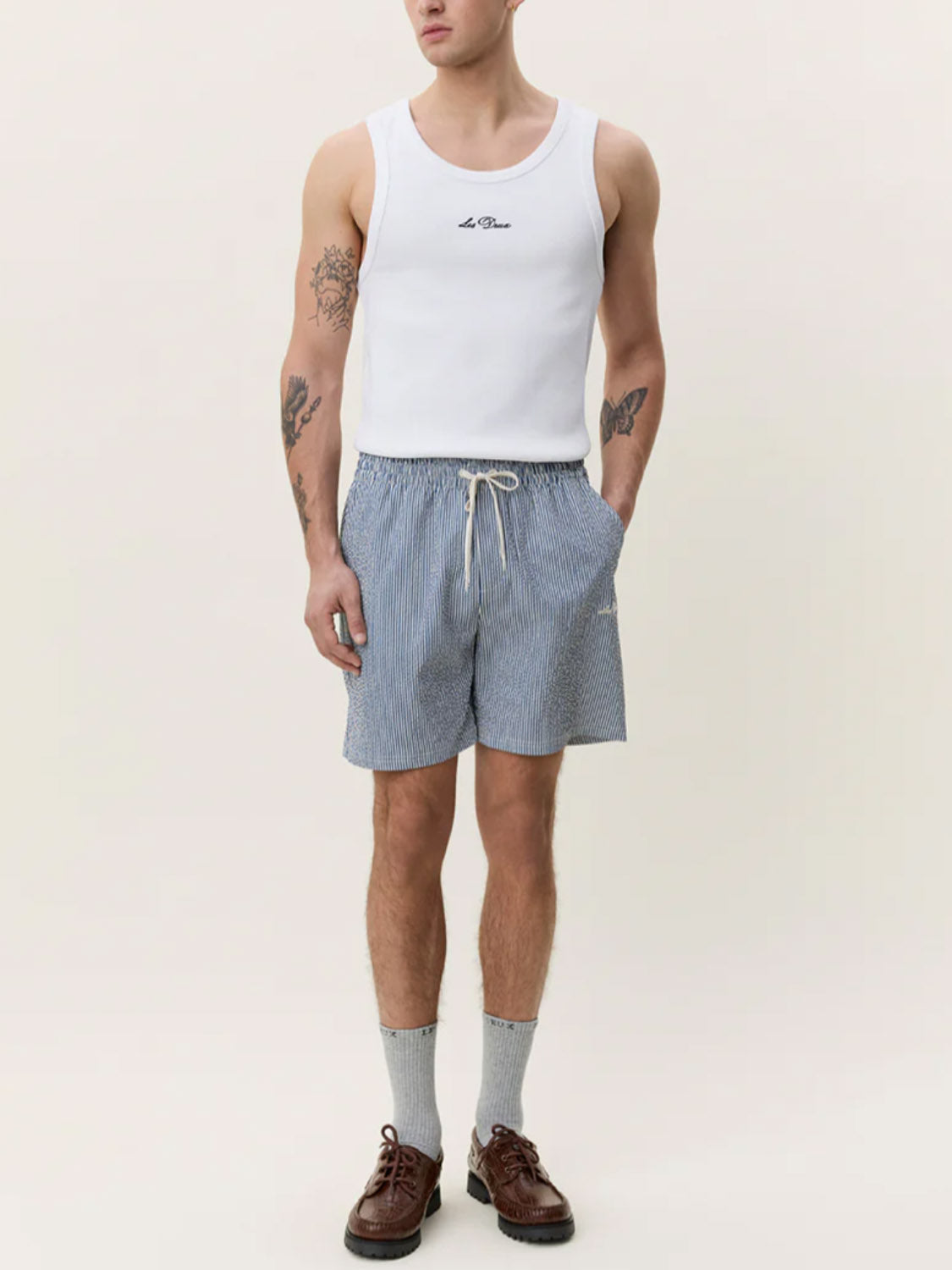 A person wears Les Deux Stripe Seersucker Swim Shorts and a white tank top, standing against a plain background. Gray socks, brown shoes, and visible tattoos on arms and legs complete their look.