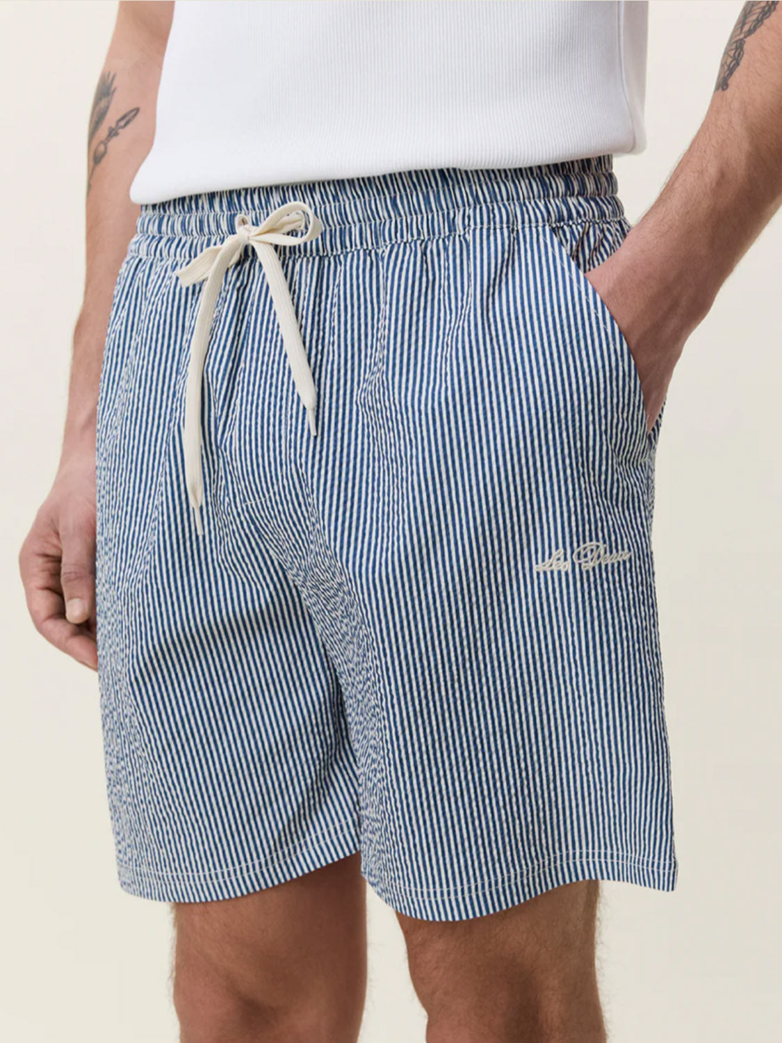 A person wears Les Deux Stripe Seersucker Swim Shorts in blue and white stripes, featuring an elastic waistband, cream drawstring, white embroidered text, and made from recycled polyester, paired with a white ribbed tank top. One hand is in the pocket.