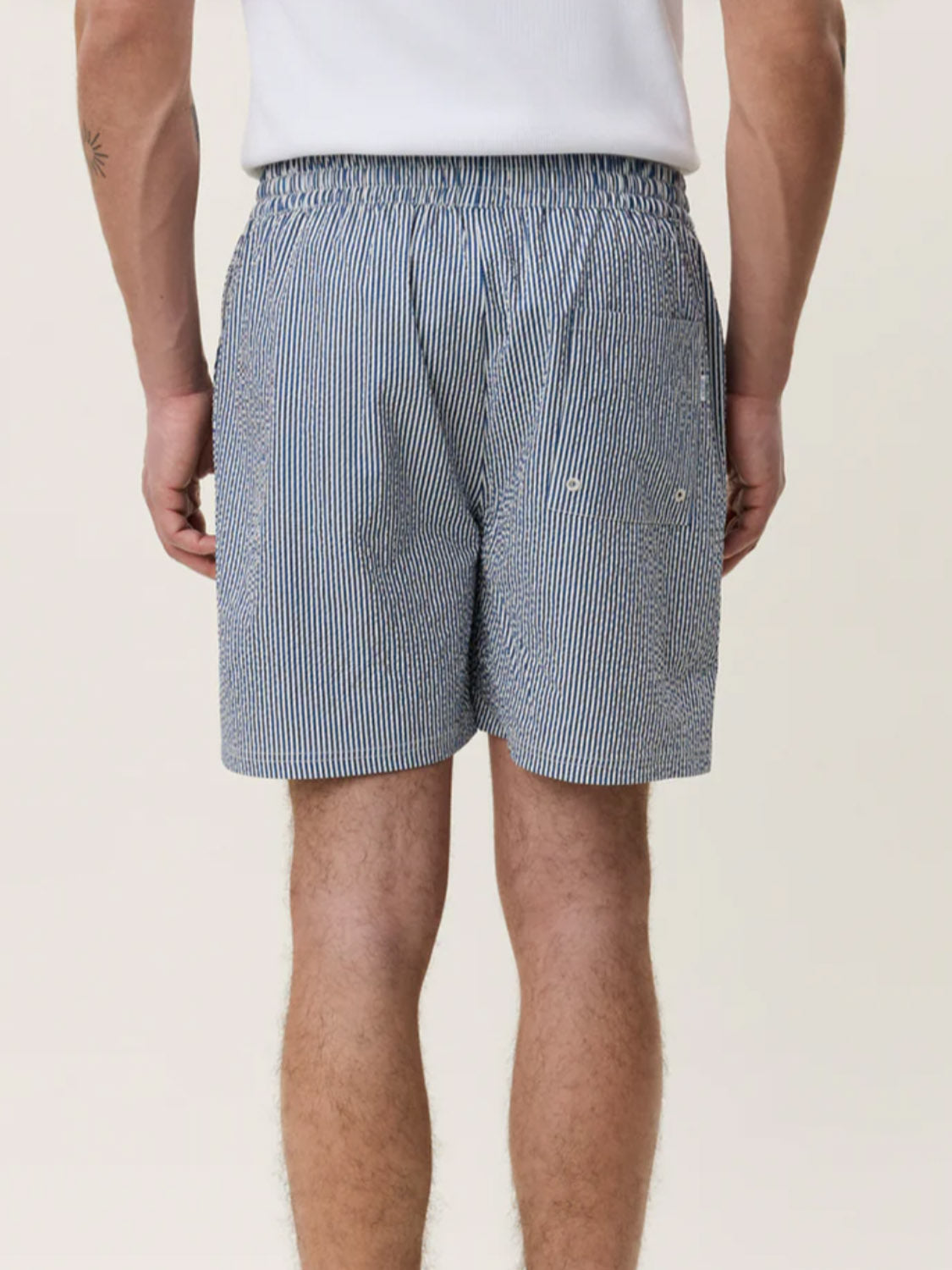 A person faces away from the camera in a white shirt and Les Deux Stripe Seersucker Swim Shorts with blue-and-white vertical stripes and an elastic waistband, set against a plain light background.