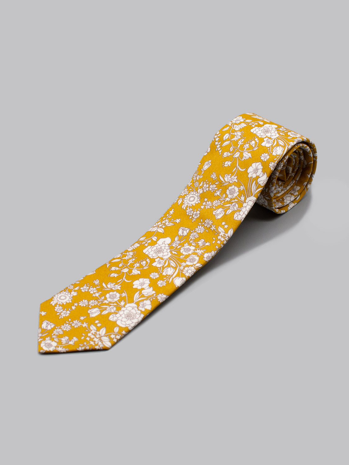 The Liberty of London Tana Lawn™ Summer tie by ID Menswear is yellow with a white floral print, handmade in Brooklyn from fine cotton. One end is rolled, and it’s displayed on a light gray background.