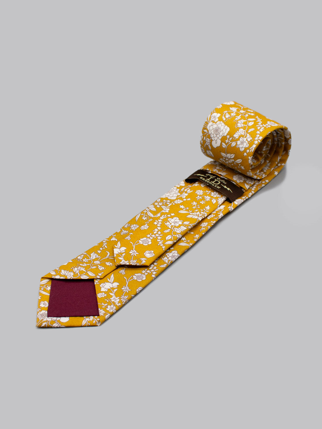 The Liberty of London Tana Lawn™ Summer tie by ID Menswear is a rolled yellow tie with a white floral pattern and maroon inner tip, handmade in Brooklyn and shown on a plain light gray background.