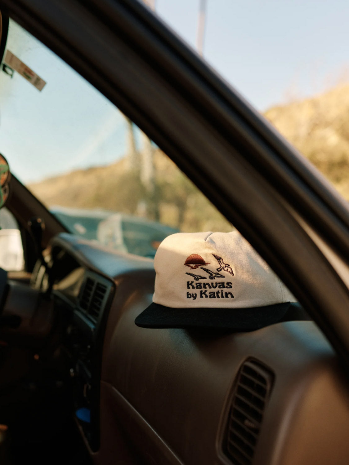 A Katin Sunset Hat with a surfing graphic sits on the cars dashboard. Made of cotton canvas, it features a snapback closure for an ideal fit. Inside the car is a brown dashboard, and outside, the landscape blurs under a clear sky.