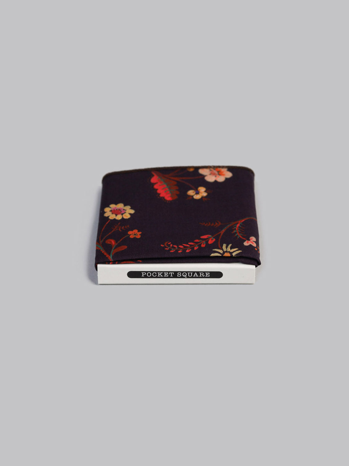 A folded Liberty™ Swedish Pocket Square by ID Menswear, featuring a red, pink, and orange floral pattern on soft Tana Lawn cotton, is shown in white POCKET SQUARE packaging against a light gray background.