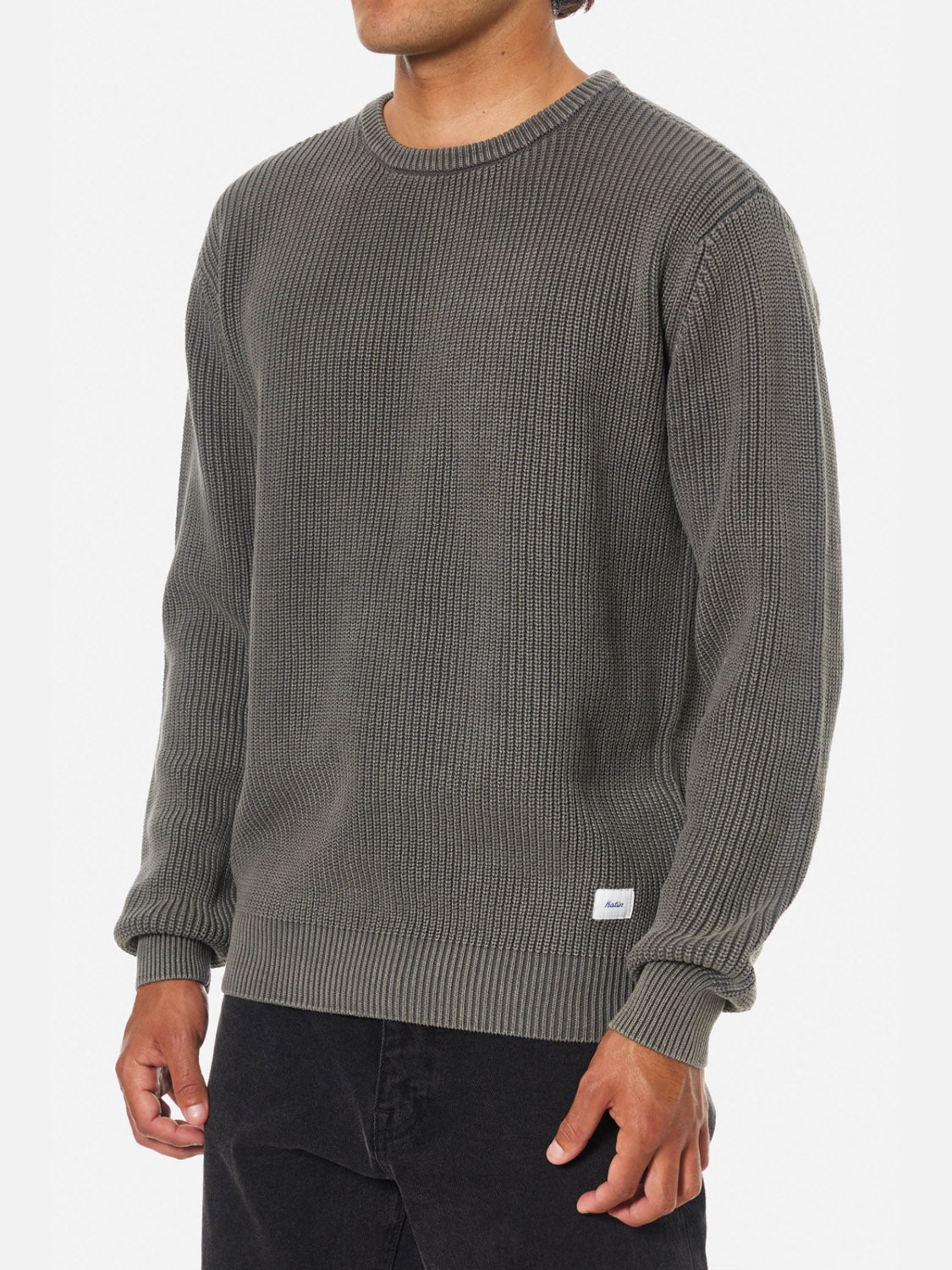 A person wears the Katin Swell Sweater—a textured, dark gray cable knit with long sleeves, ribbed pattern, vintage wash finish, and a small white tag near the hem—paired with black pants. The person's head is not visible.