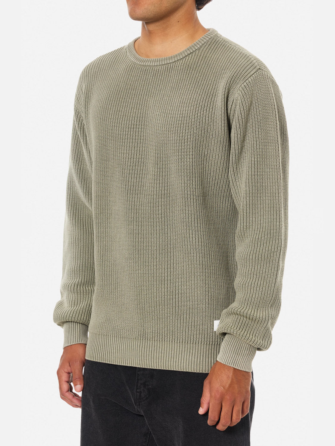 A person, head partially out of frame, stands against a white background wearing the Katin Swell Sweater in light olive green (100% cotton, textured knit, long sleeves) paired with black pants.
