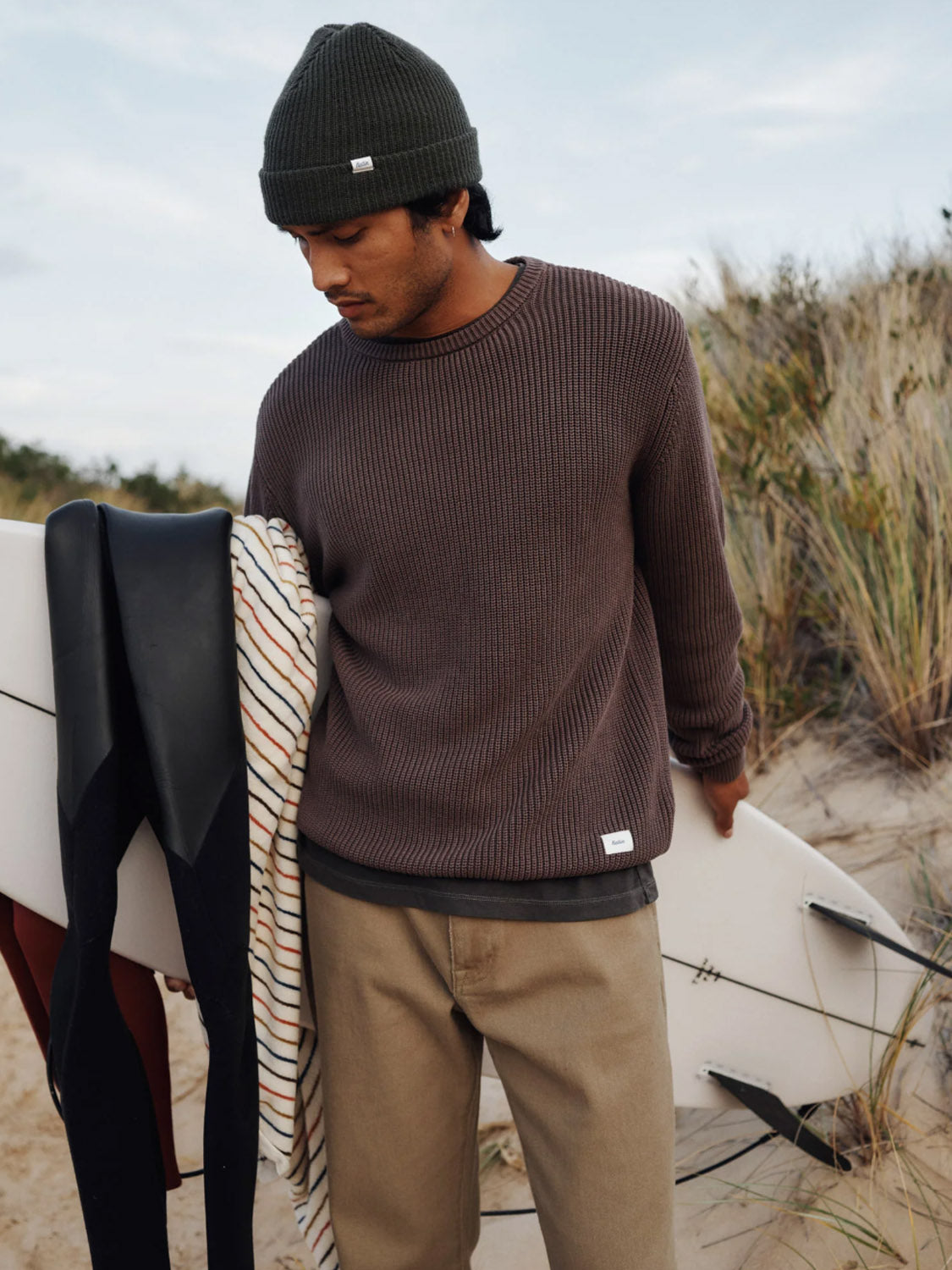 A man in a dark knit beanie, Katin Swell Sweater, and khaki pants stands on a sandy beach with a surfboard and striped towel, tall grass and dunes behind him.