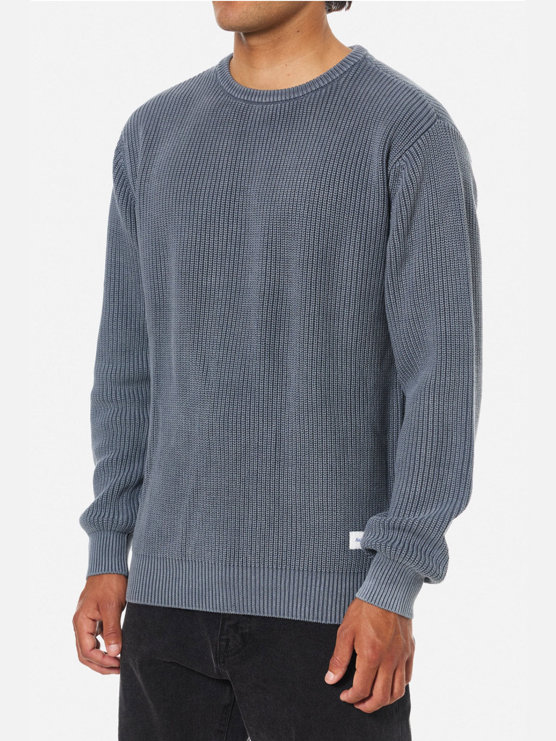 A person models the Katin Swell Sweater, a blue cable knit with long sleeves and a relaxed fit, paired with black pants against a plain white background. The person's head is partially out of frame in the image.
