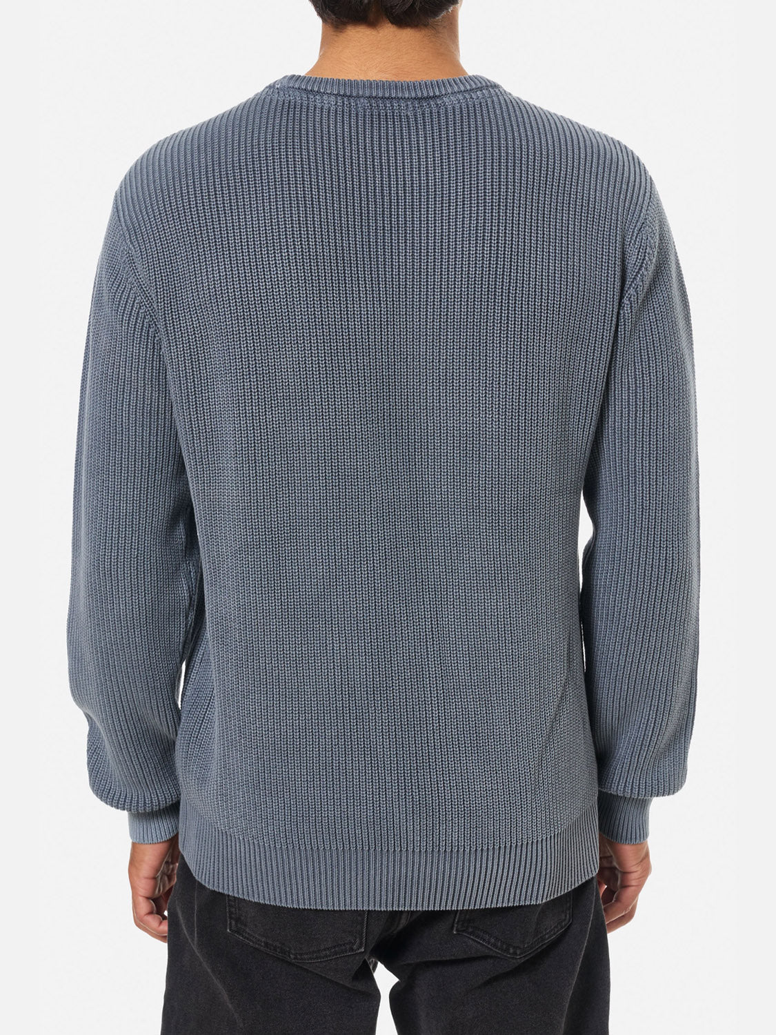 A person is shown from the back wearing the Katin Swell Sweater, a relaxed fit, long-sleeved blue cable knit paired with black pants. The background is plain white.