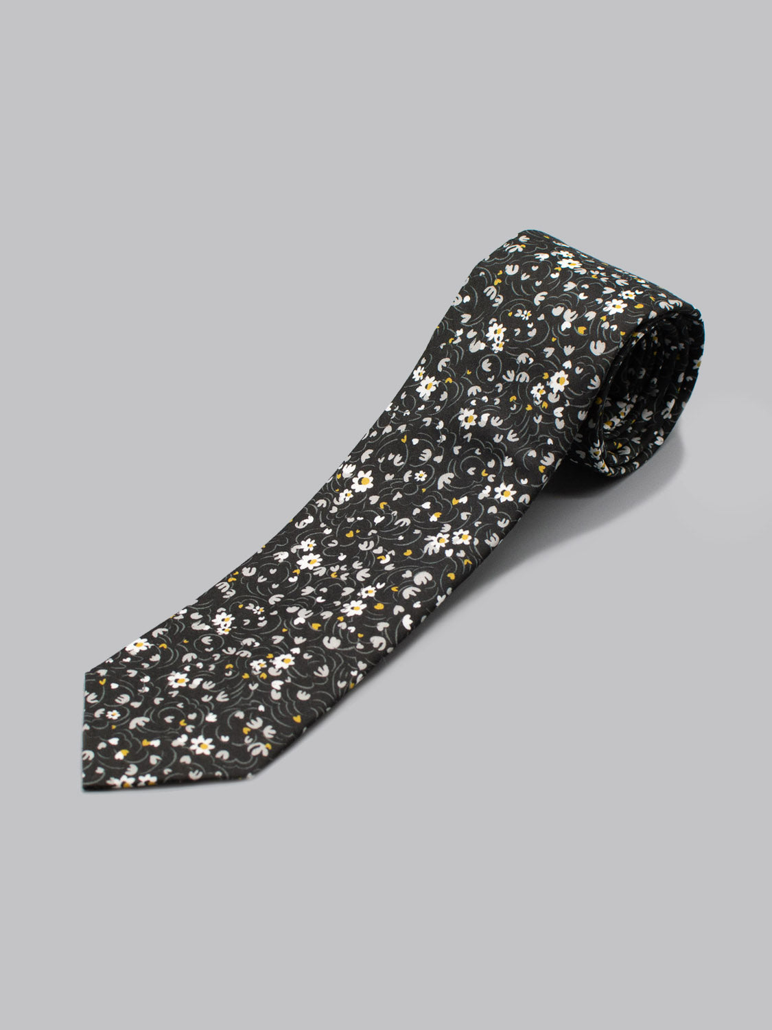 The ID Menswear Liberty of London Tana Lawn™ Swirl Tie is handmade in Brooklyn from soft cotton, featuring a black base with small white and yellow flowers and green leaves on a light gray background.