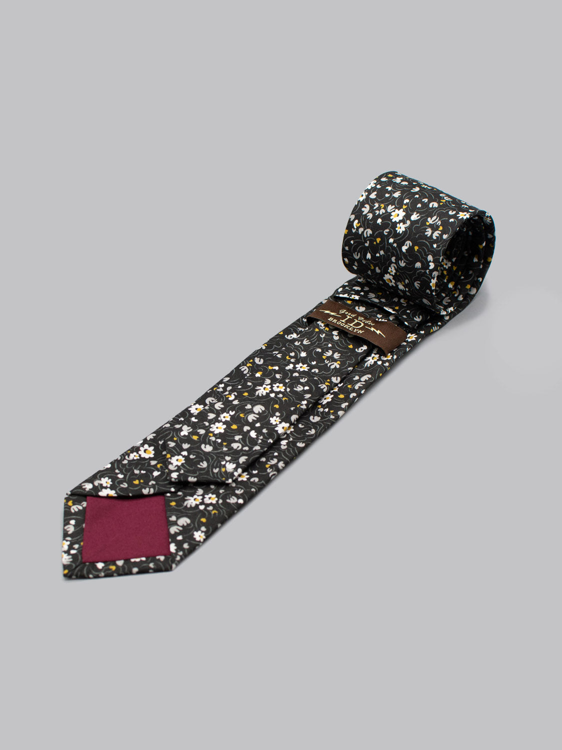 The Liberty of London Tana Lawn™ Swirl Tie by ID Menswear is a black tie with a small white and yellow floral pattern, unrolled on a gray background. Handmade in Brooklyn, its tip features maroon accent lining on the back.