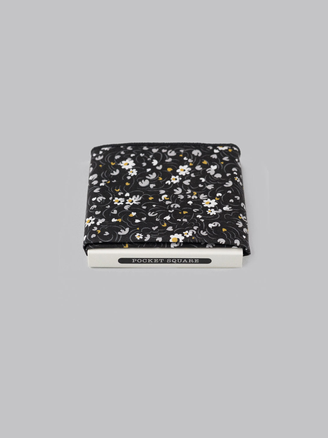A folded black Liberty™ Swirl Pocket Square by ID Menswear, featuring a white and yellow floral design in Tana Lawn cotton, peeks from a silver holder marked POCKET SQUARE against a plain gray background.