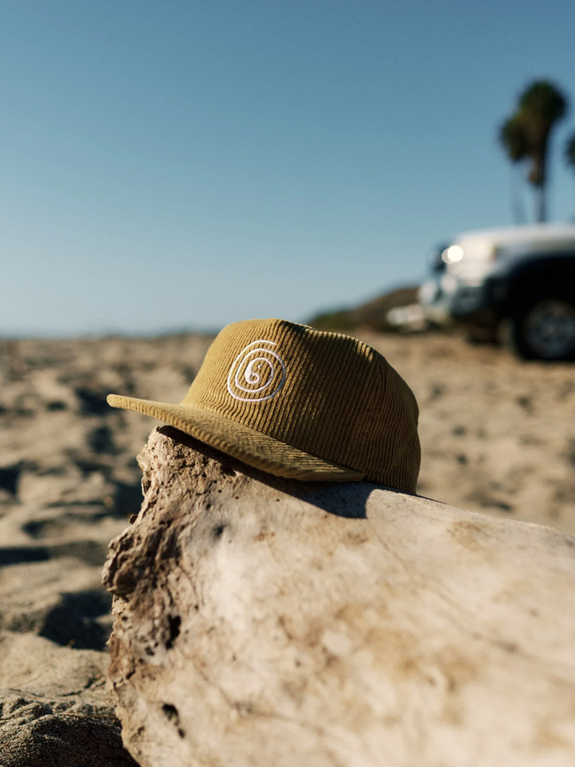 A tan Katin Swirl Hat, featuring a white logo, sits on weathered driftwood on a sandy beach. In the backdrop, blurred palm trees and a vehicle are visible beneath the clear blue sky.