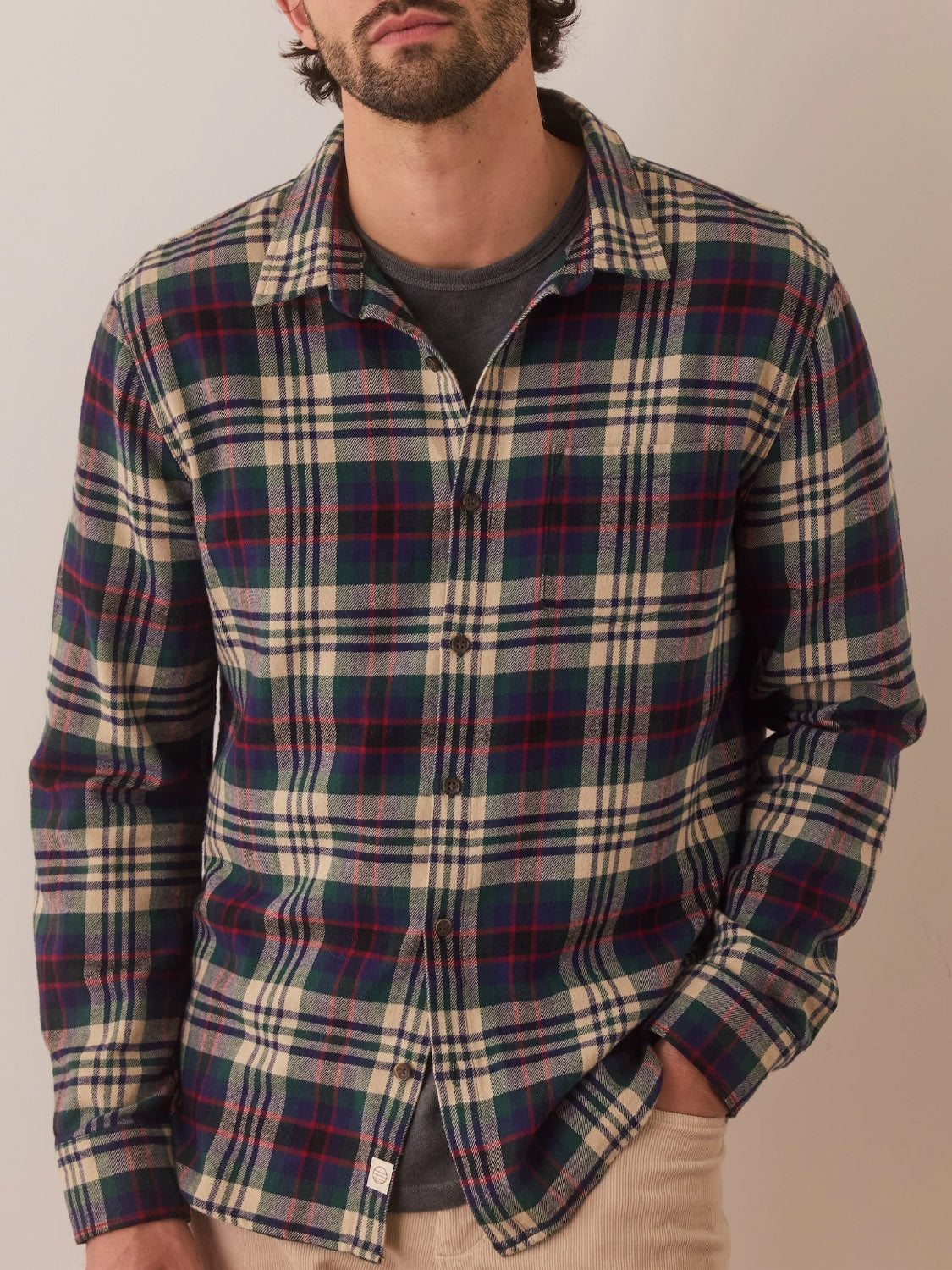 A person in a Marine Layer Tahoe Flannel Shirt, featuring a blue, red, and white plaid pattern stands against a neutral background with one hand in their pocket. The 100% cotton fabric provides brushed softness, complemented by a dark undershirt and light beige pants for timeless style.