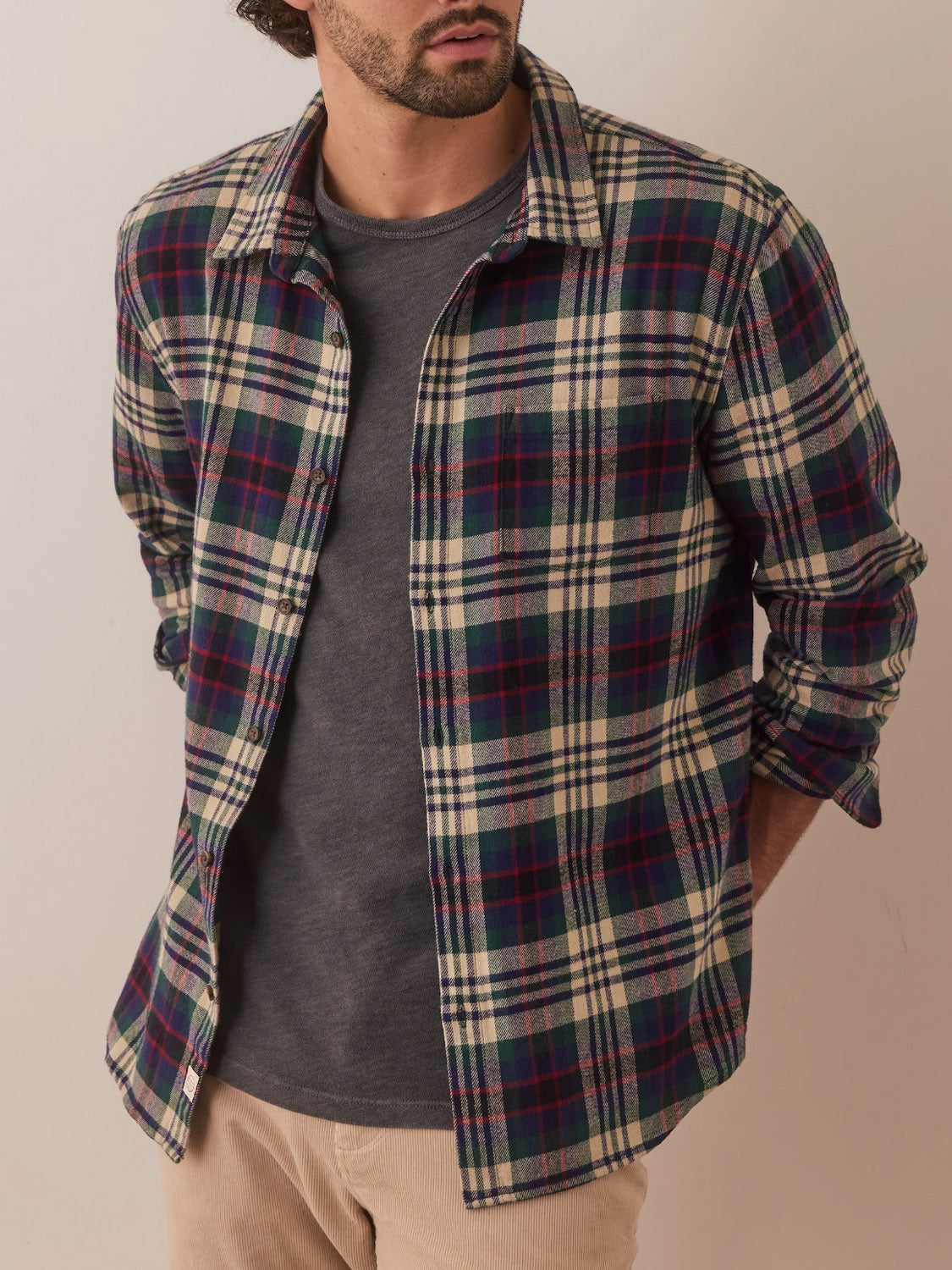 A man stands against a light-colored wall in the Marine Layer Tahoe Flannel Shirt, featuring red, green, and cream plaid. His gray t-shirt and beige pants complement the 100% cotton flannels brushed softness as his hands rest casually in his pockets.