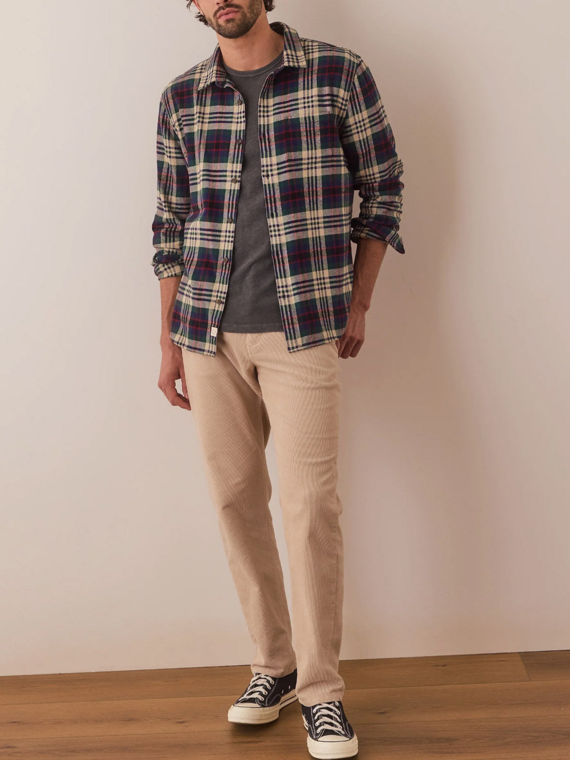 A man leans casually against a wall on a wooden floor, wearing the Marine Layer Tahoe Flannel Shirt famous for its brushed softness over a dark t-shirt. His beige pants complement his black sneakers. With one hand in his pocket and the other resting by his side, he exudes relaxed confidence.