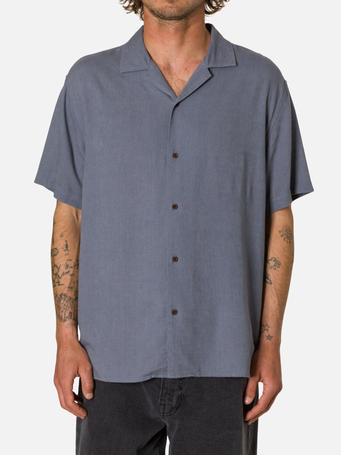 Someone wears the Katin Teller Shirt— a short-sleeve, camp-collar, button-up gray rayon linen shirt—untucked over dark pants, with tattooed arms visible. The background is plain white.