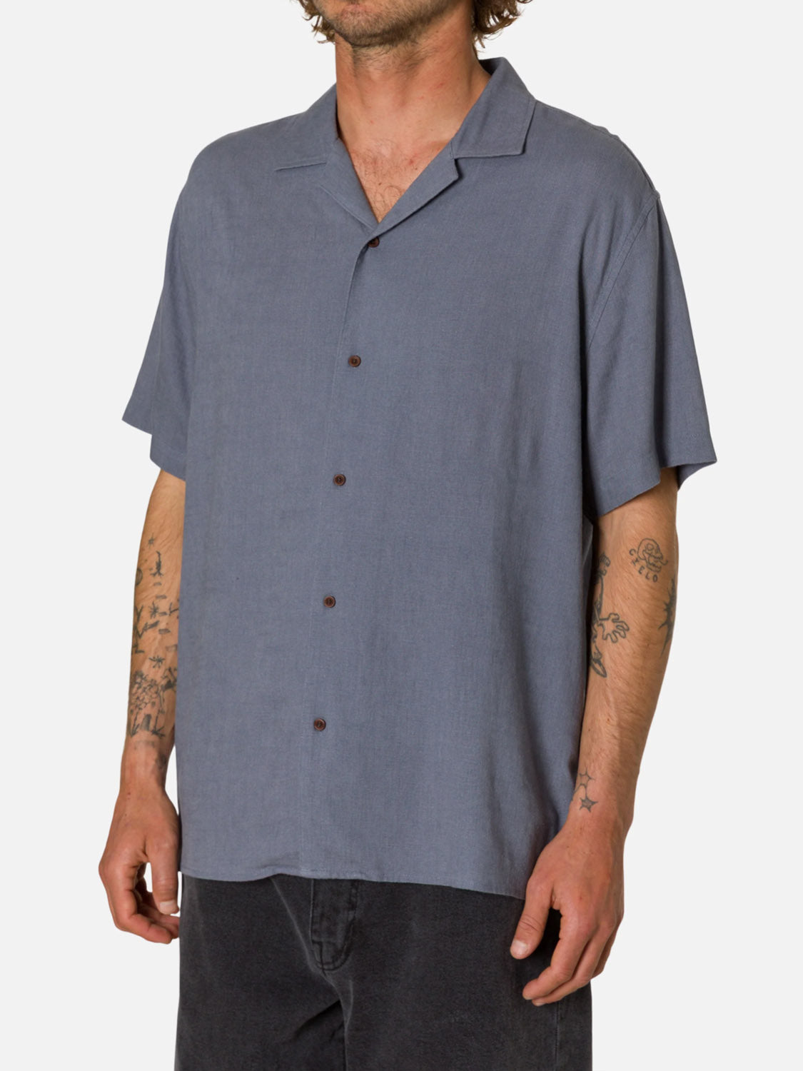 A man stands against a plain white background wearing the Katin Teller Shirt—a light gray-blue, short-sleeved camp collar button-up with slub texture—paired with dark pants and displaying tattoos on his forearms.