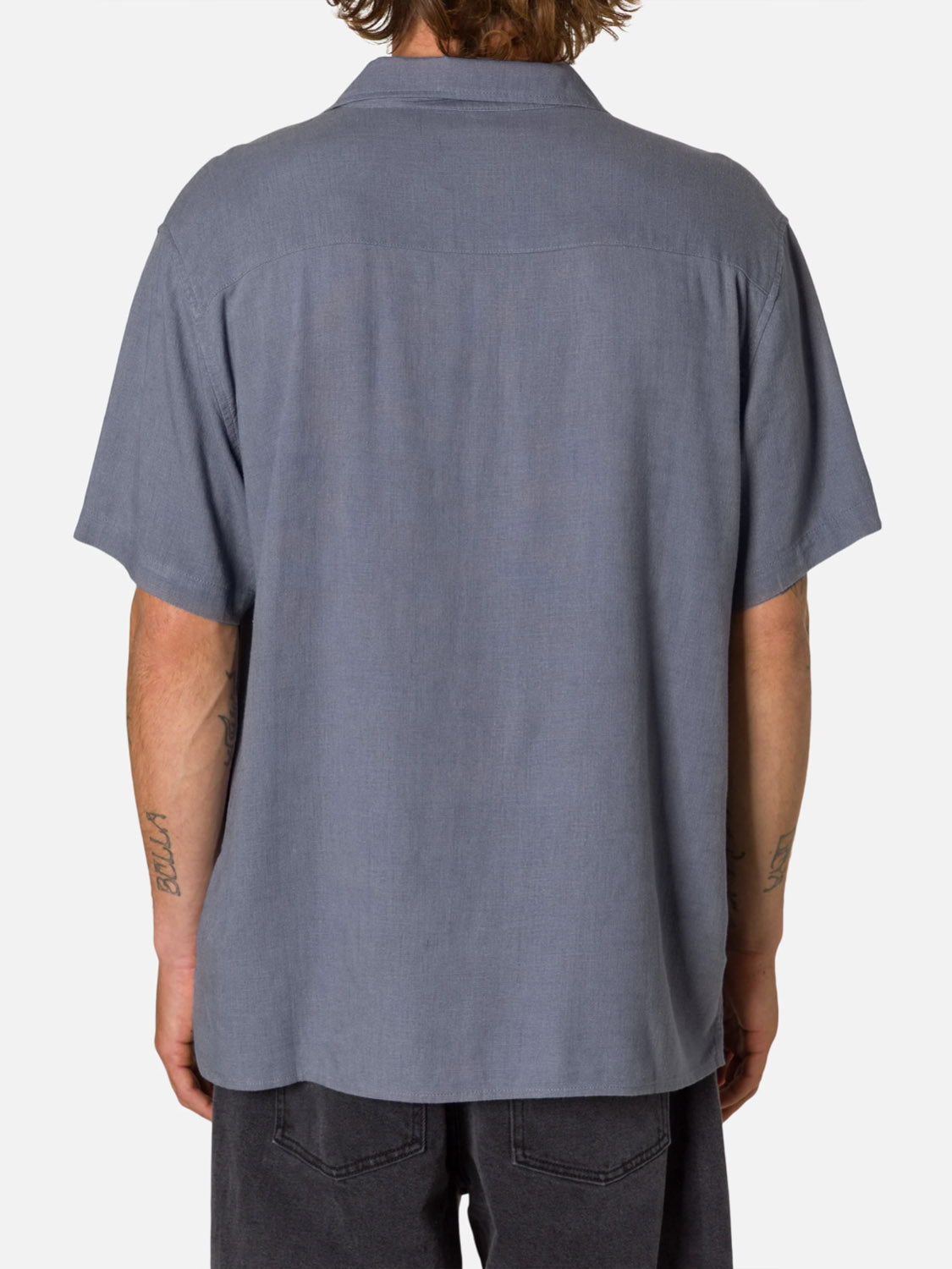 A tattooed person is seen from behind wearing the Katin Teller Shirt, a light blue rayon linen camp collar style, paired with dark pants against a plain white background.