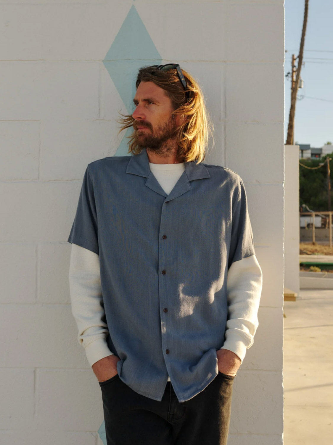 A man with long blond hair and a beard leans against a white brick wall, sunglasses on his head, wearing the Katin Teller Shirt over a white long-sleeve shirt with dark pants, hands in his pockets.