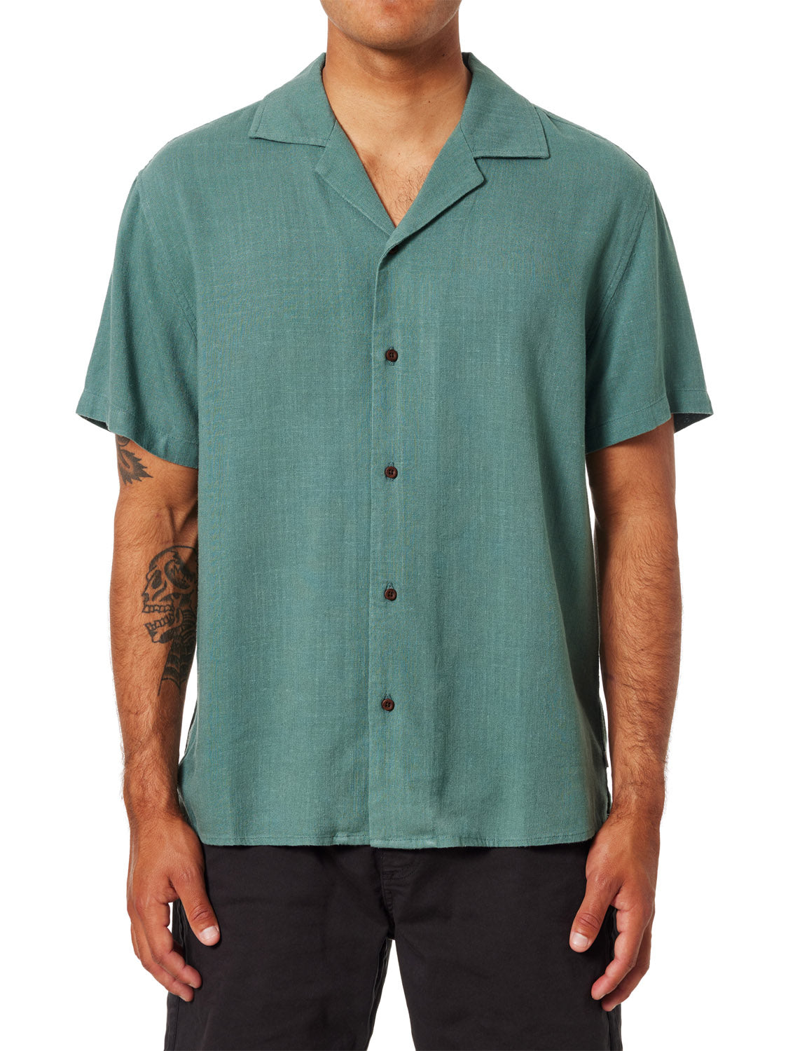 A person is wearing a Katin Teller Shirt, short-sleeved and green, with a camp collar over black pants. This rayon linen blend shirt has a relaxed design and a visible tattoo of a skull on their right arm against the plain white background.