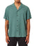 A person is wearing a Katin Teller Shirt, short-sleeved and green, with a camp collar over black pants. This rayon linen blend shirt has a relaxed design and a visible tattoo of a skull on their right arm against the plain white background.