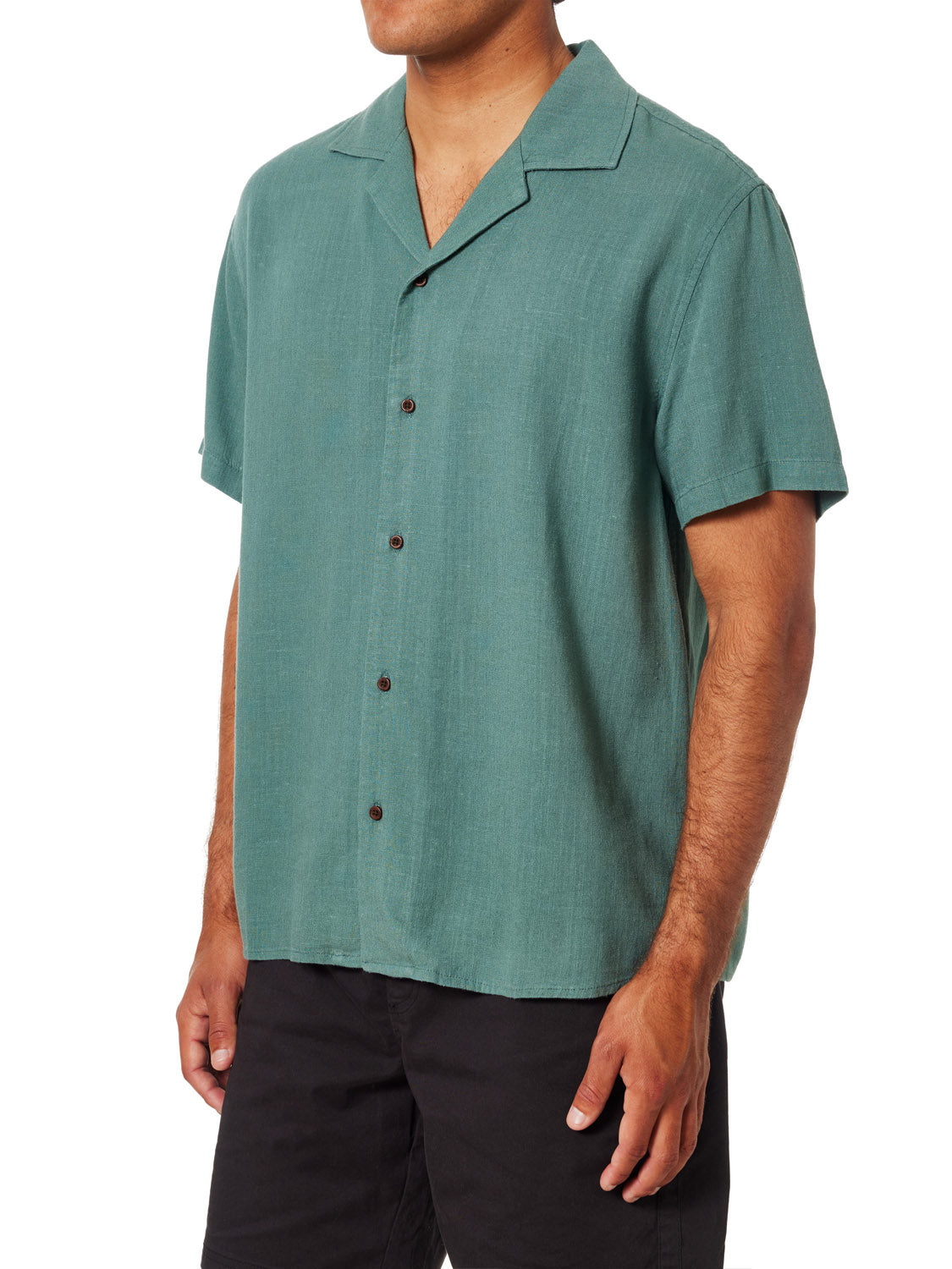 A person in a green Katin Teller Shirt, made from a rayon linen blend, stands against a white background. The shirt features short sleeves, a camp collar, and dark buttons and is paired with black pants. Their relaxed hands contribute to the casual elegance of the ensemble.