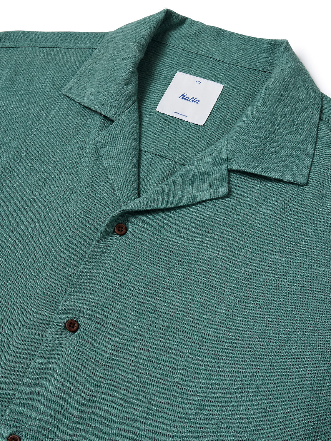 A close-up of the Katin Teller Shirt reveals a green hue with dark buttons and a neck label. Made from a rayon linen blend, it boasts a textured look and chic camp collar.