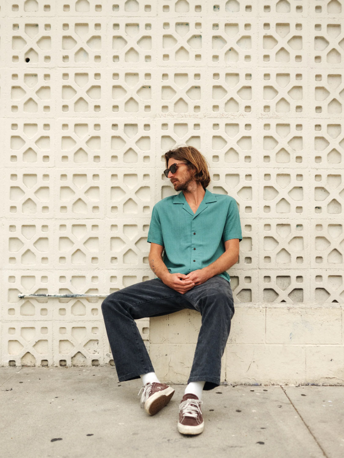 A man with shoulder-length hair and sunglasses, wearing a teal Katin Teller Shirt made of a rayon linen blend, sits relaxed against a patterned white wall. He looks sideways, one leg stretched out in black jeans and brown sneakers.