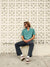 A man with shoulder-length hair and sunglasses, wearing a teal Katin Teller Shirt made of a rayon linen blend, sits relaxed against a patterned white wall. He looks sideways, one leg stretched out in black jeans and brown sneakers.