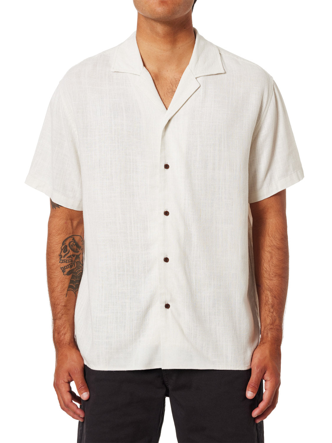 Someone is wearing a white, short-sleeved Katin Teller Shirt featuring a camp collar and seven dark buttons, paired with black pants. A tattoo is visible on their right forearm, but their face is not shown.
