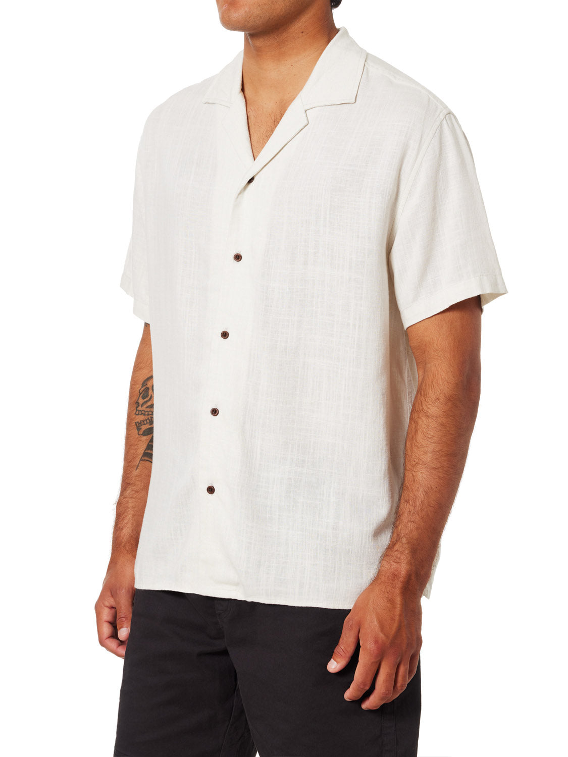 A person is wearing a short-sleeve, white Katin Teller Shirt with textured pattern and dark buttons. Made from a rayon-linen blend, it has a camp collar for style. The outfit is completed with black pants and a visible forearm tattoo, while the arms are relaxed at their sides.