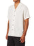 A person is wearing a short-sleeve, white Katin Teller Shirt with textured pattern and dark buttons. Made from a rayon-linen blend, it has a camp collar for style. The outfit is completed with black pants and a visible forearm tattoo, while the arms are relaxed at their sides.