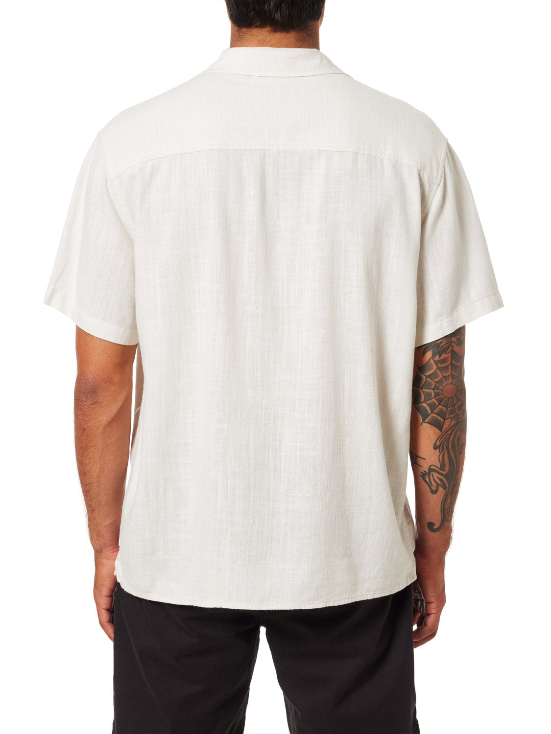 A person in a light beige Katin Teller Shirt, made from rayon linen blend and featuring a camp collar, is seen from the back. Their right arm displays an intricate sleeve tattoo, paired elegantly with sleek black pants.