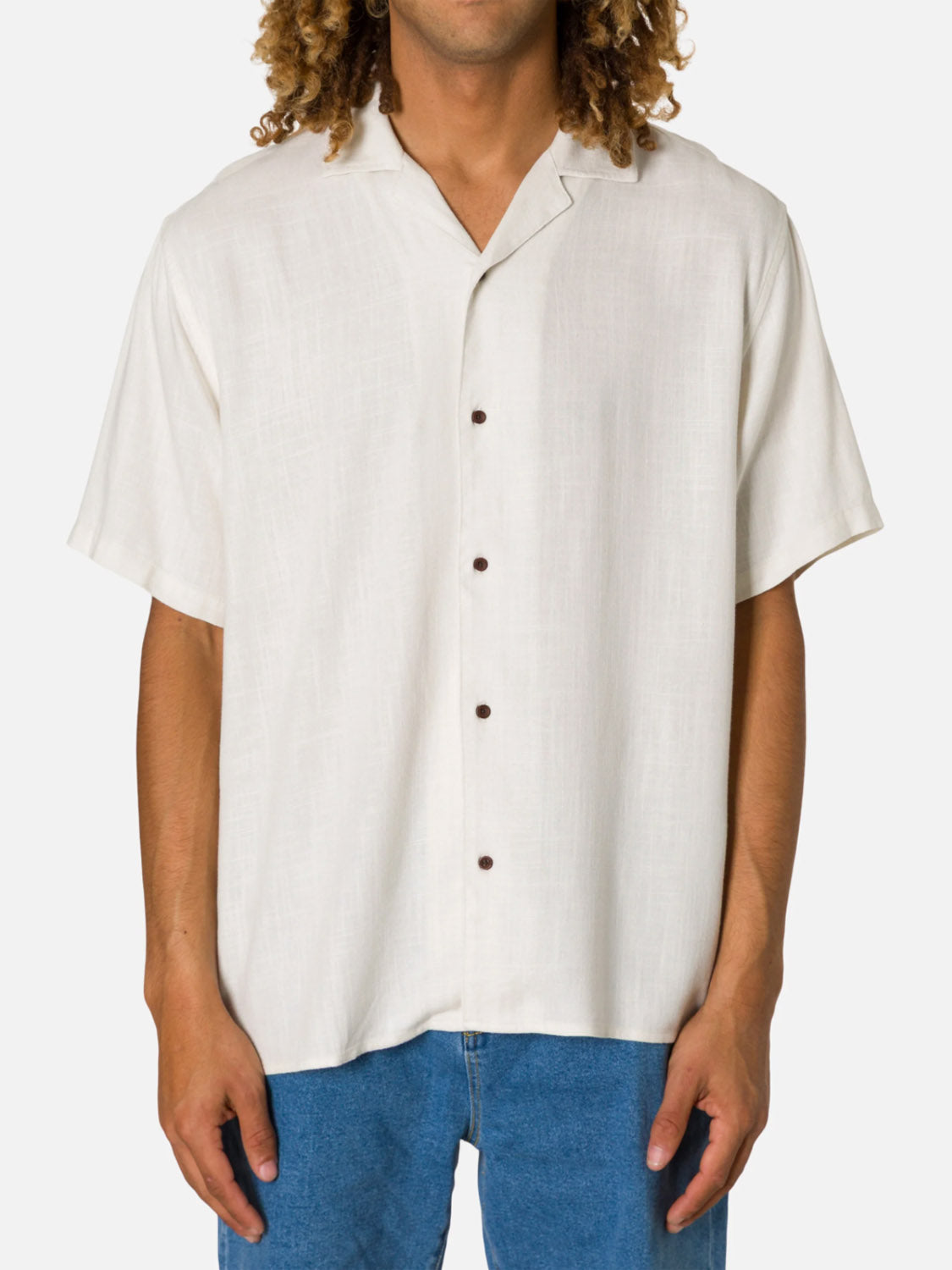 A person stands against a plain white background wearing the Katin Teller Shirt—a short-sleeve, button-up white camp collar shirt with brown buttons and slub texture—and blue jeans. Their head is partially out of frame.