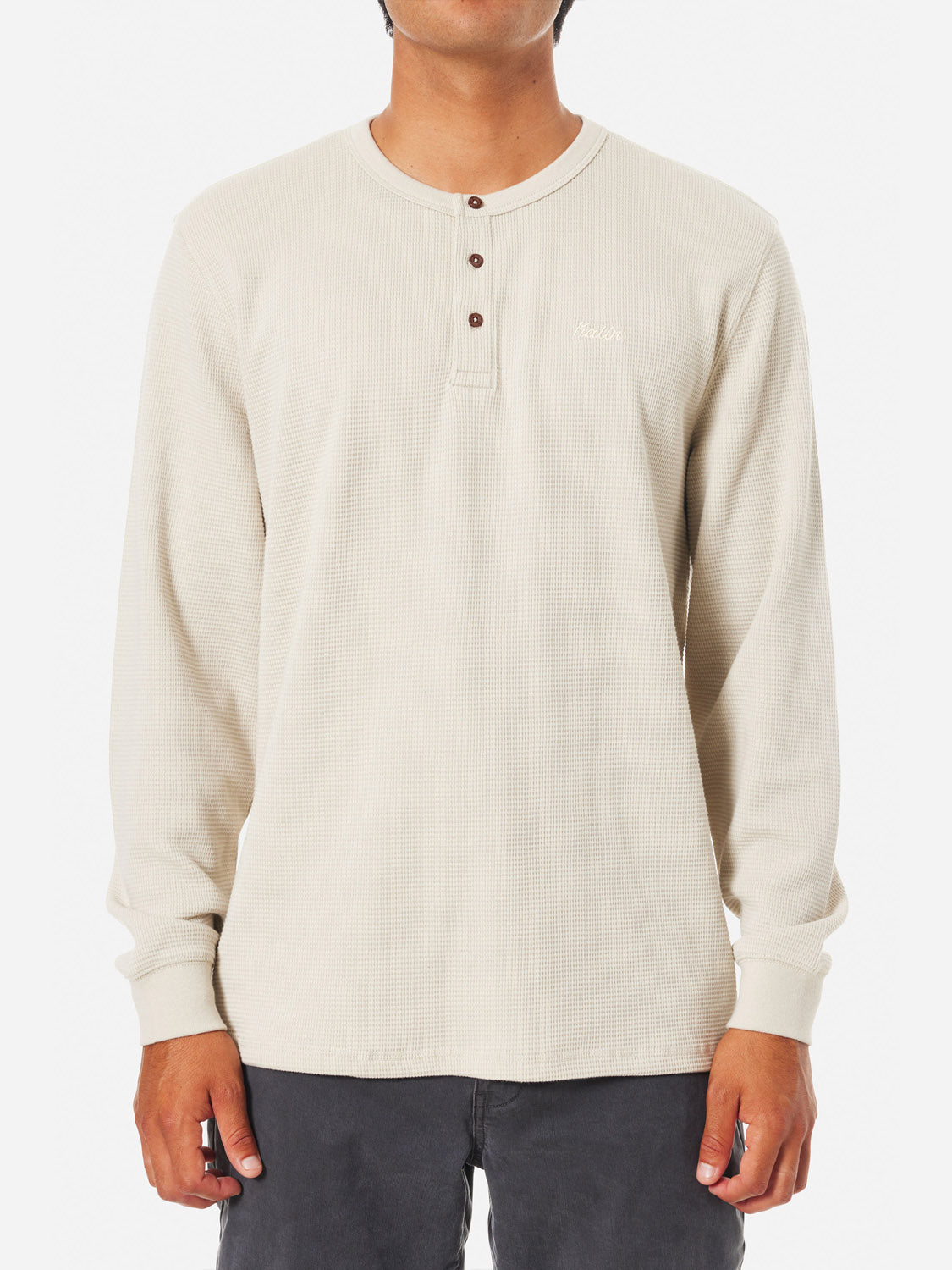 Against a white background, a person wears the Katin Mesa Thermal, a beige waffle knit Henley by Katin. Featuring three buttons and an embroidered logo on the left chest, this cotton-polyester blend shirt pairs effortlessly with dark pants.