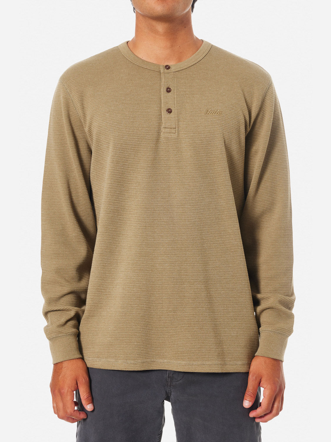 A person is wearing the Katin Mesa Thermal in light brown, featuring a 3-button henley design and a subtle chest logo. Crafted from a soft cotton-polyester blend, its paired with dark pants against a plain white background.