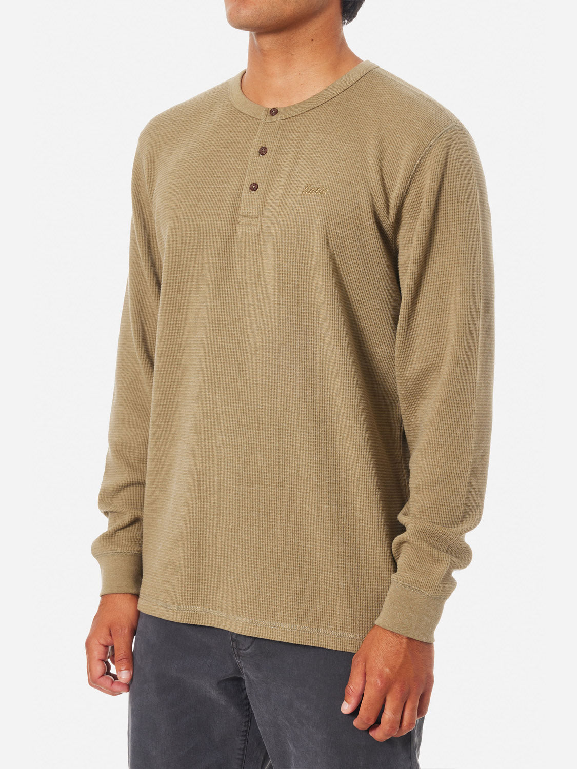 A person wears the Katin Mesa Thermal, an olive green Henley shirt made from a cotton-polyester blend with a subtle textured fabric. Paired with dark pants, the side view from shoulder to mid-thigh stands out vividly against a white background.